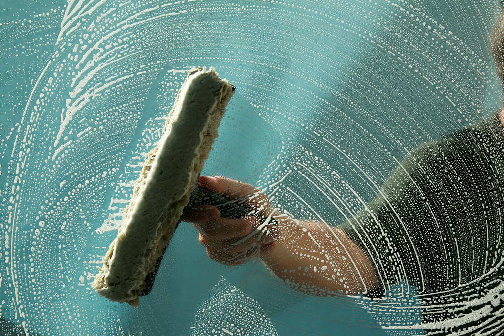 Professional window cleaner spreading white soap suds across a glass window pane with a washer sleeve — residential window cleaning, Christchurch NZ