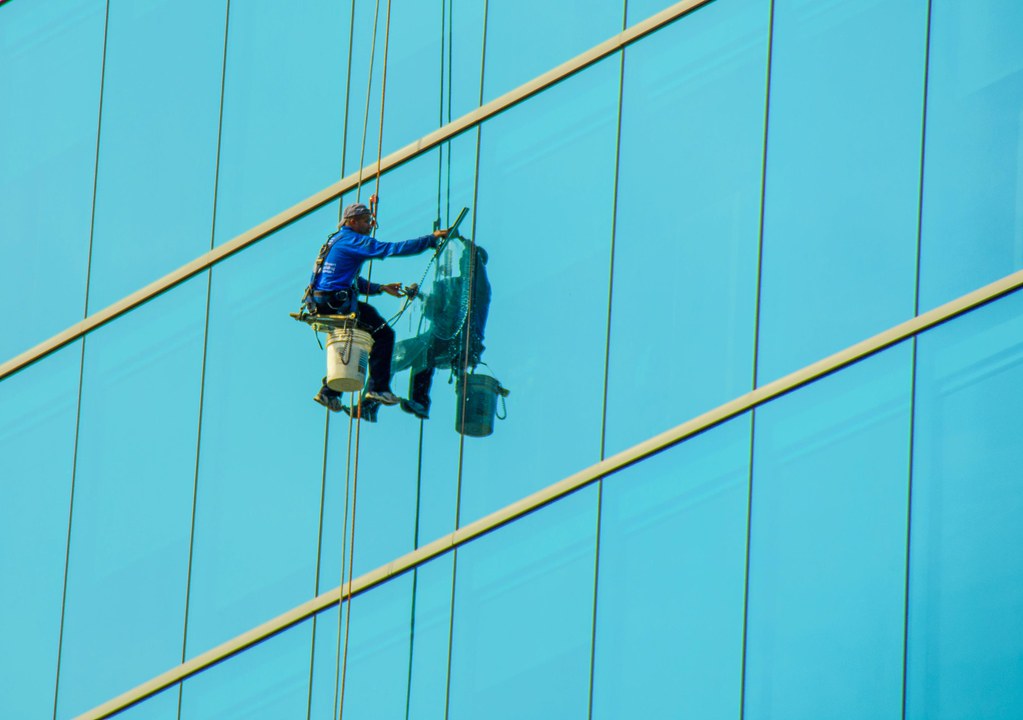 Rope-access window cleaner with bucket squeegeeing a blue high-rise glass facade — high-rise window cleaning