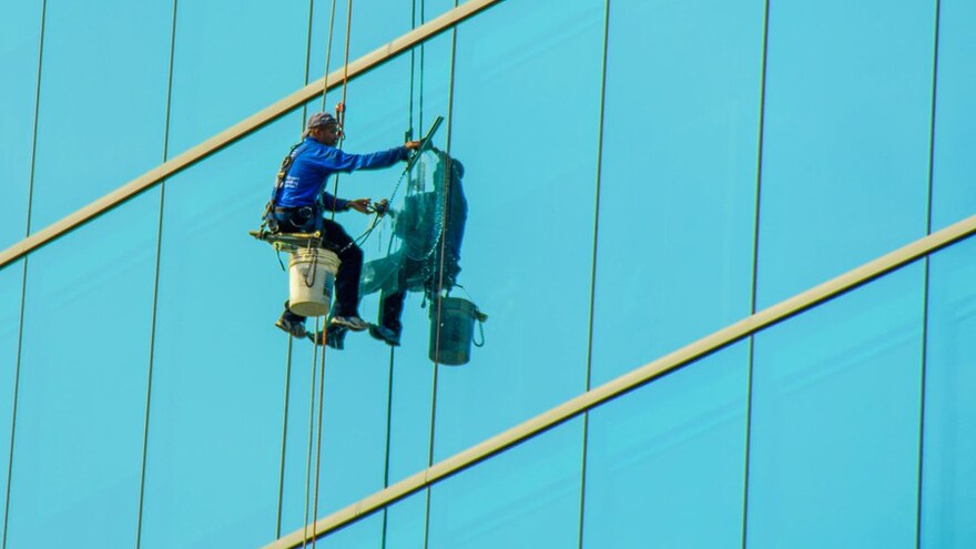 Rope-access window cleaner with bucket squeegeeing a blue high-rise glass facade — high-rise window cleaning